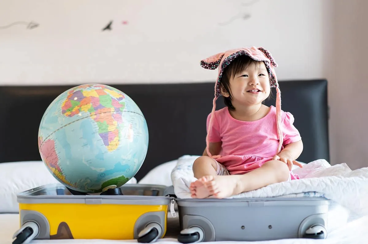 An open suitcase is on the bed, a toddler sits in one half smiling and there's a globe in the other.