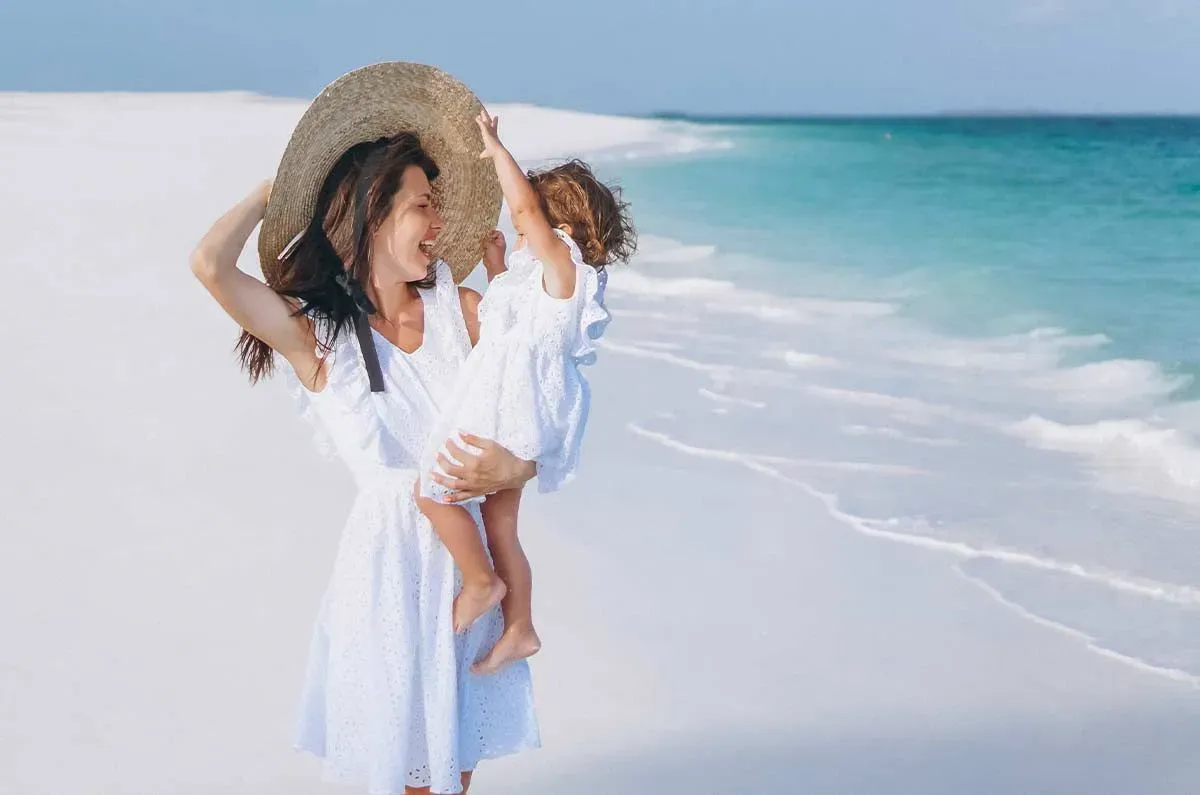 Mum smiling while she holds her toddler in her arms on the beach.