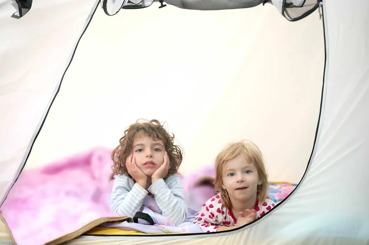 Two toddlers lying on their stomachs in their camping tent looking out the zip door.