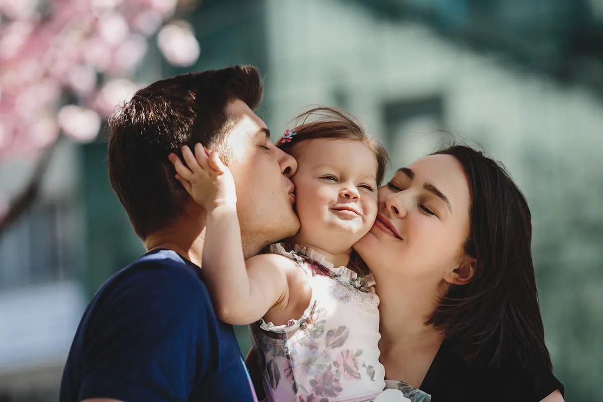 Parents holding their young daughter and kissing her on either cheek.