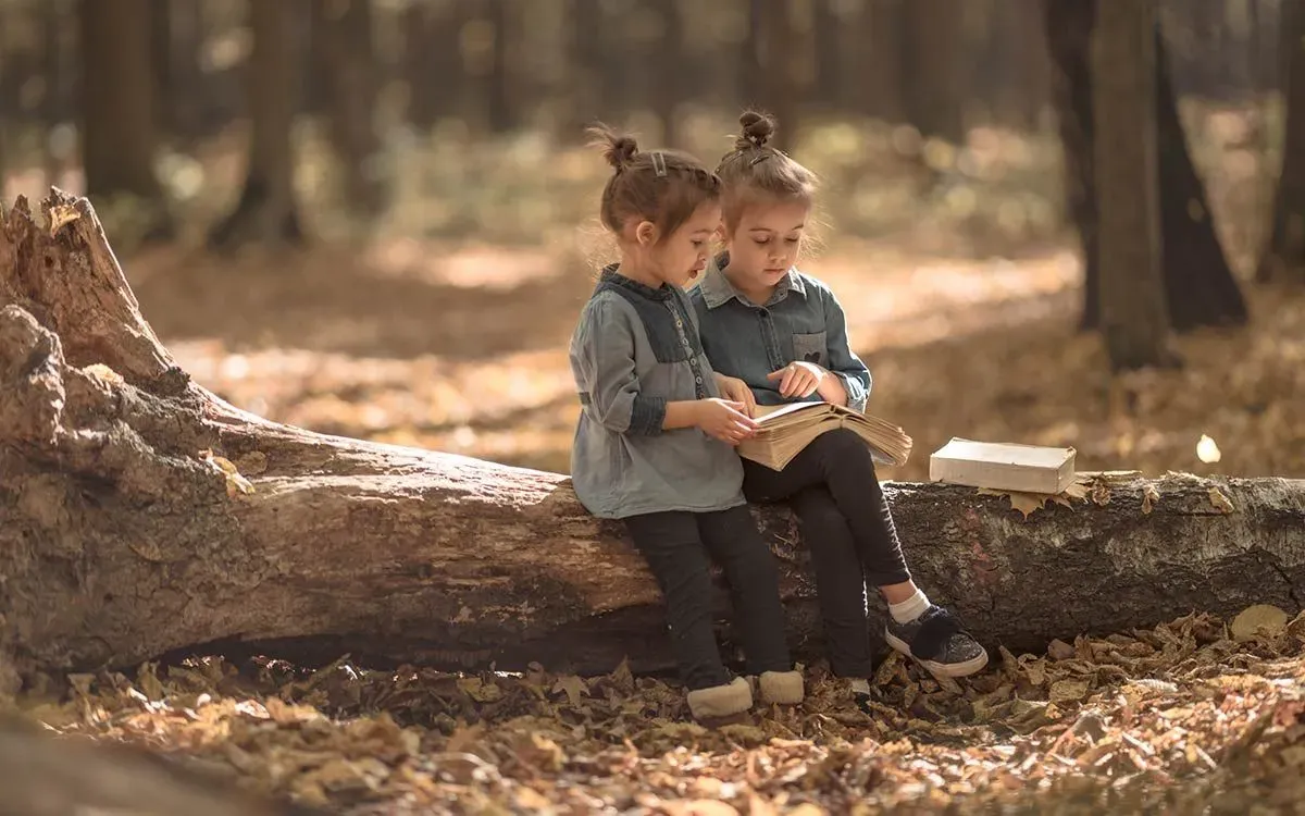 Two little girls sat outside on a tree trunk reading a book together for their book review.