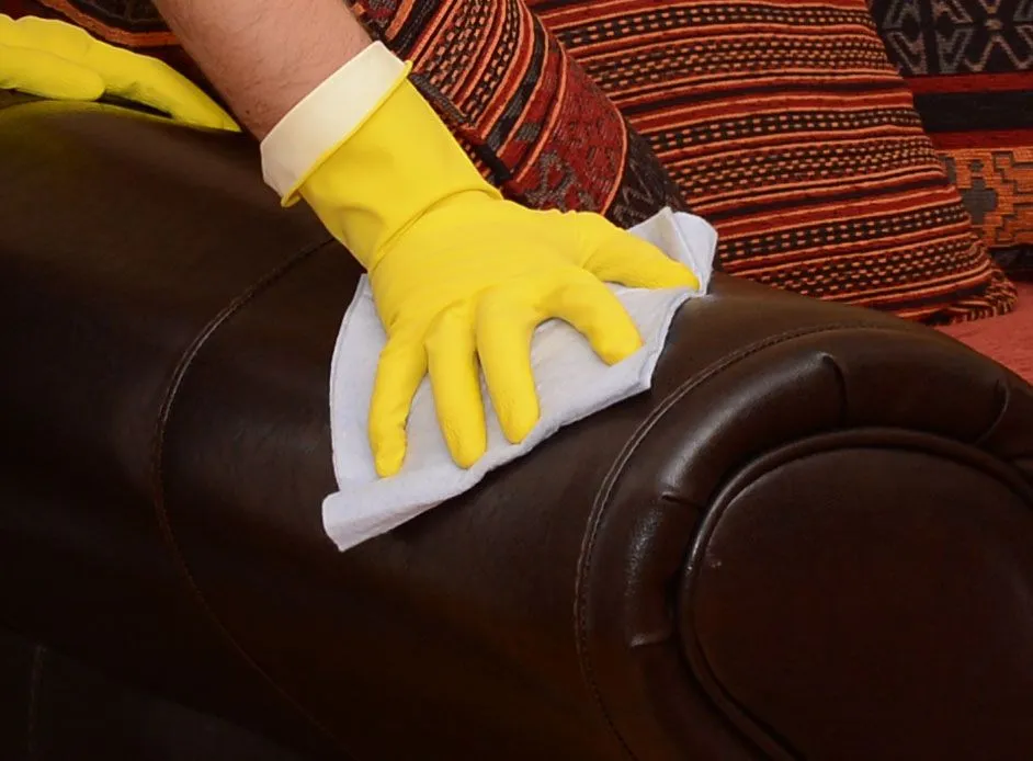 A close up image of a gloved hand rubbing a brown leather sofa with a cloth in an attempt to remove a biro stain.