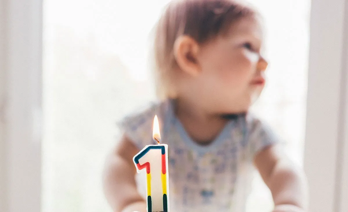 A toddler sits behind a cake with a colourful lit candle in the shape of the number one.