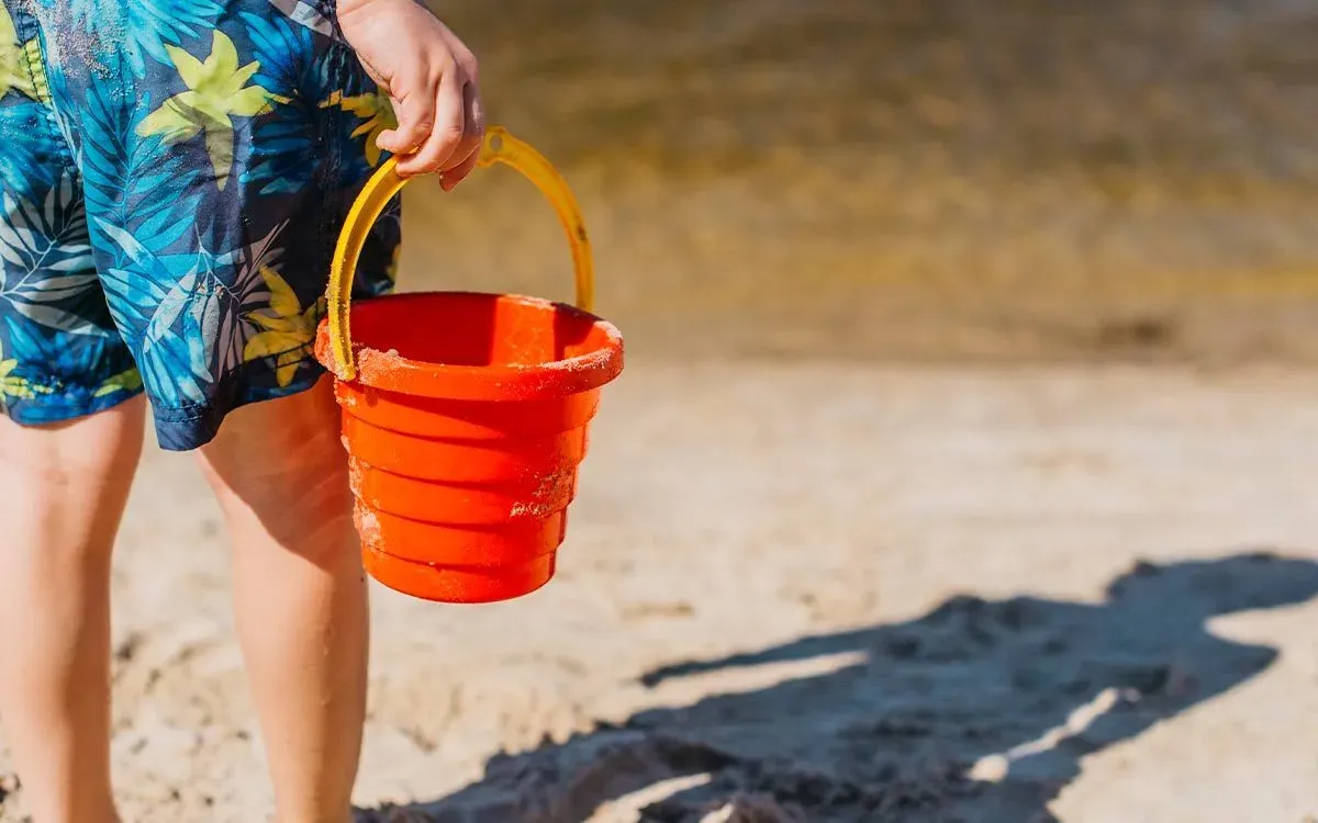 Boy standing on the beach holding a bucket, his shadow is in the sand in front of him.