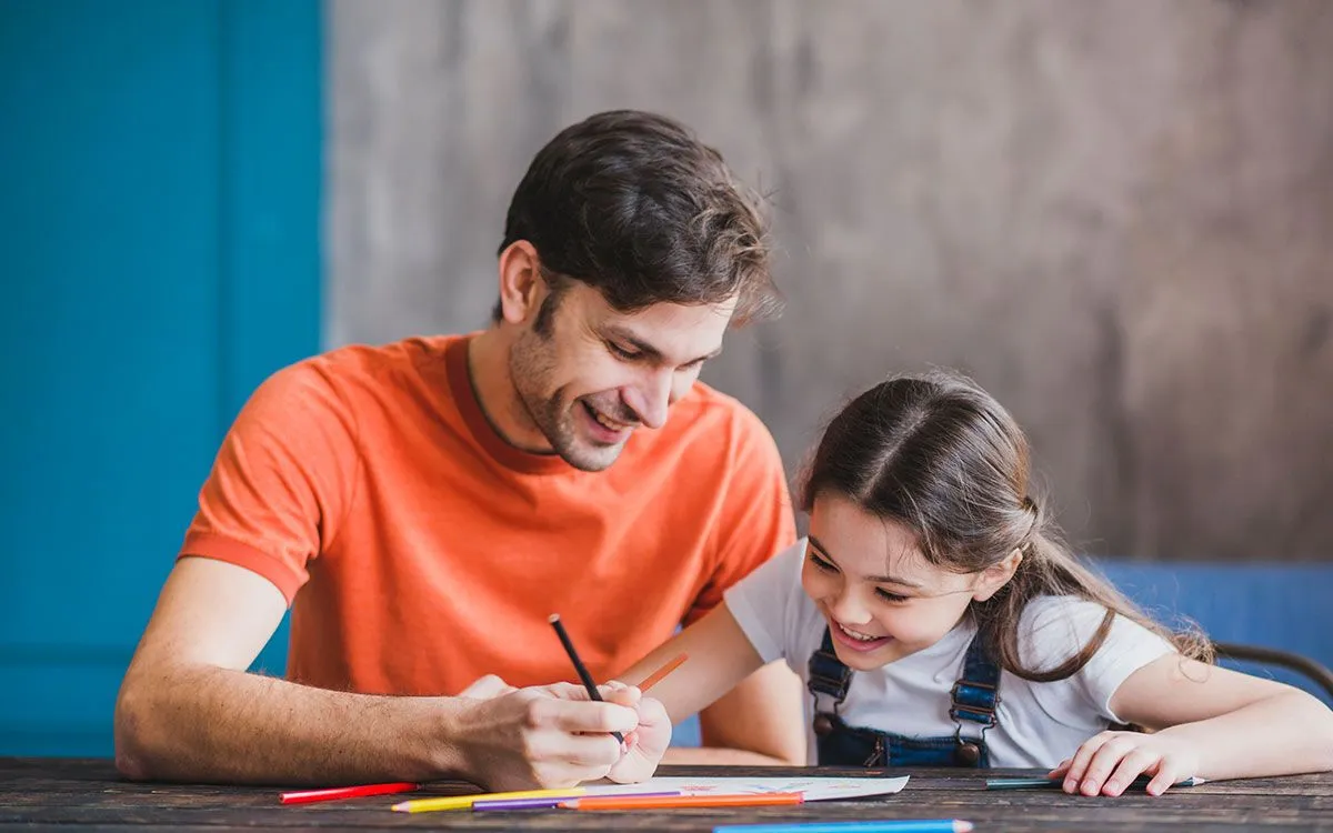 A father and daughter sit together at the table with pens and paper, they are learning about the present perfect tense.