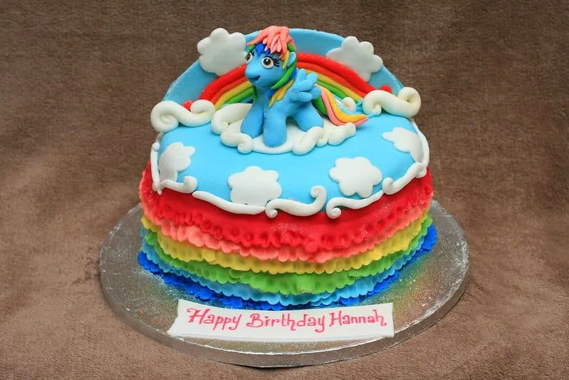 A My Little Pony Cake with blue icing on top, rainbow coloured icing stripes around the side and a My Little Pony character (Rainbow Dash) cake topper sitting on top. 