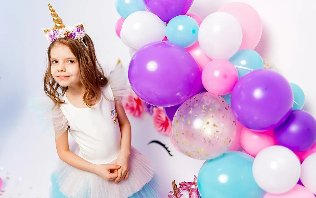 A young girl is dressed as a pony for a My Little Pony birthday party, she is stood next to a purple, pink, white and blue balloon display.