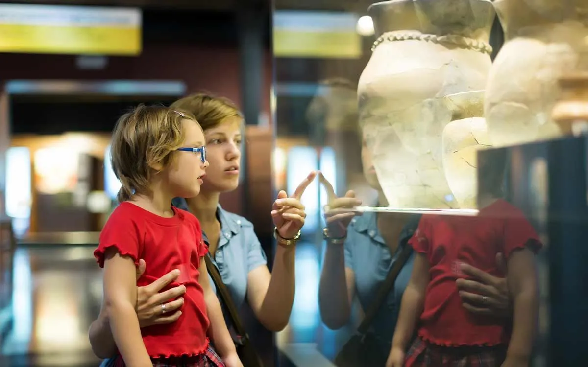 Mum pointing out Ancient Greek artefacts in a display cabinet in a museum to her daughter.