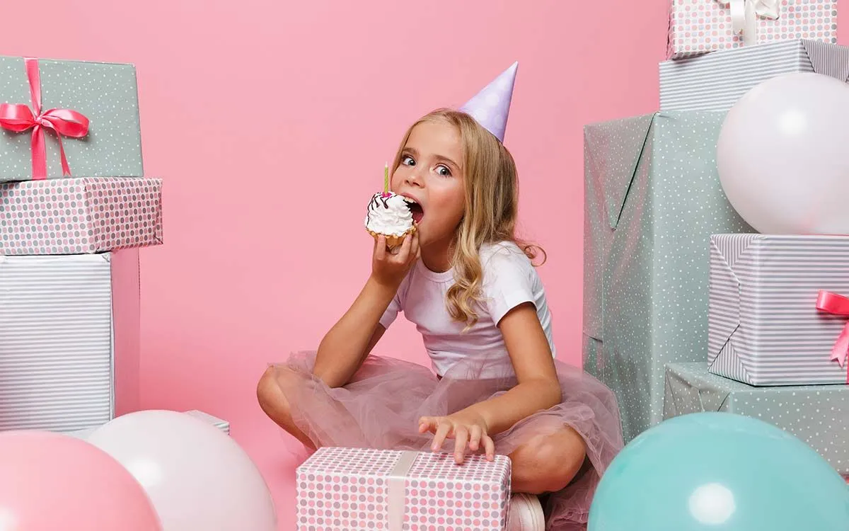 A young girl looks into the camera as she takes a bite of a cupcake, she is wearing a party hat and is surrounded by presents and balloons.