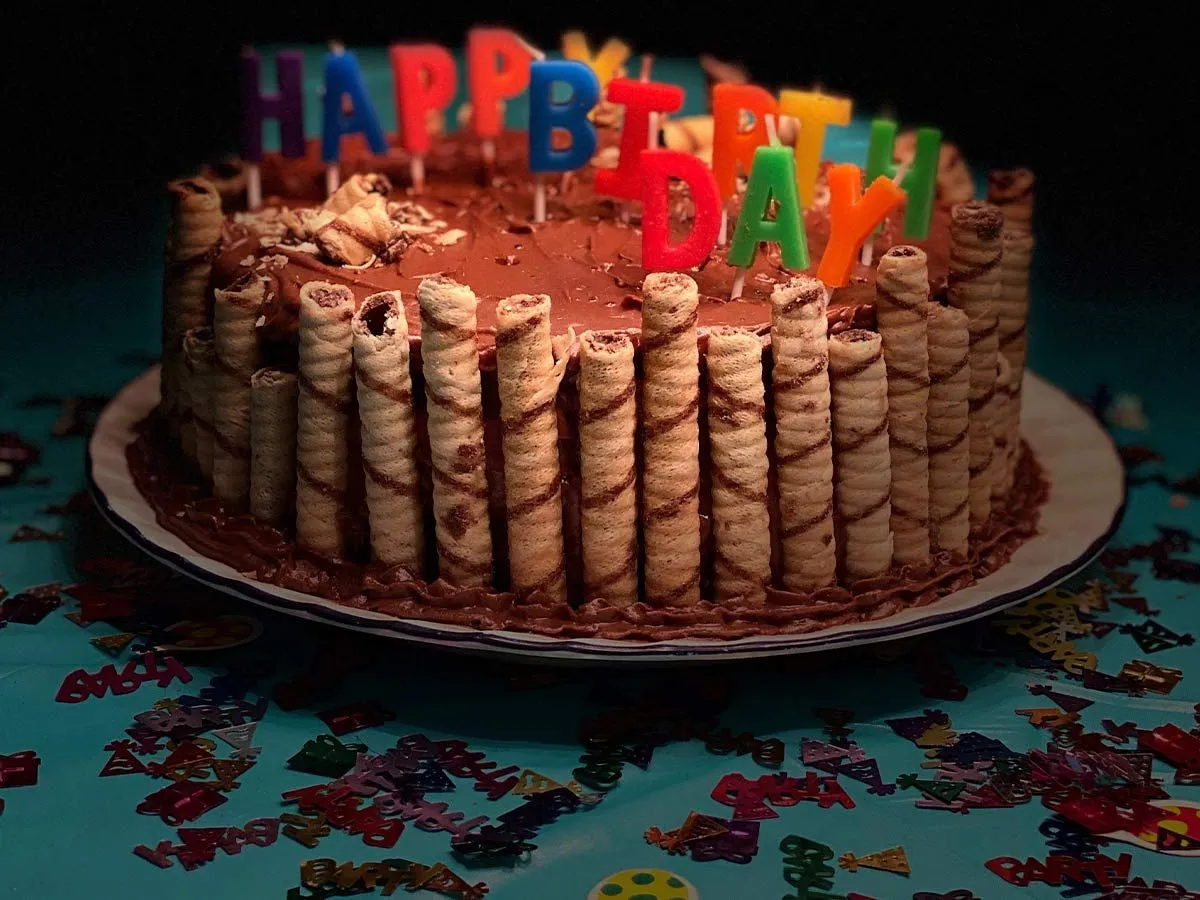 A chocolate cake topped with chocolate icing, with wafers rolls around the side and candles spelling 'happy birthday' on top.
