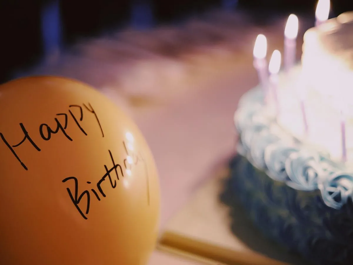 A yellow balloon with 'happy birthday' written on it, in the background is a birthday cake with lit candles.