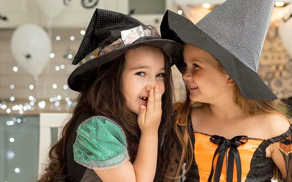 Two young girls dressed in Halloween witch costumes, one smiles at her friend whilst the other looks at the camera.