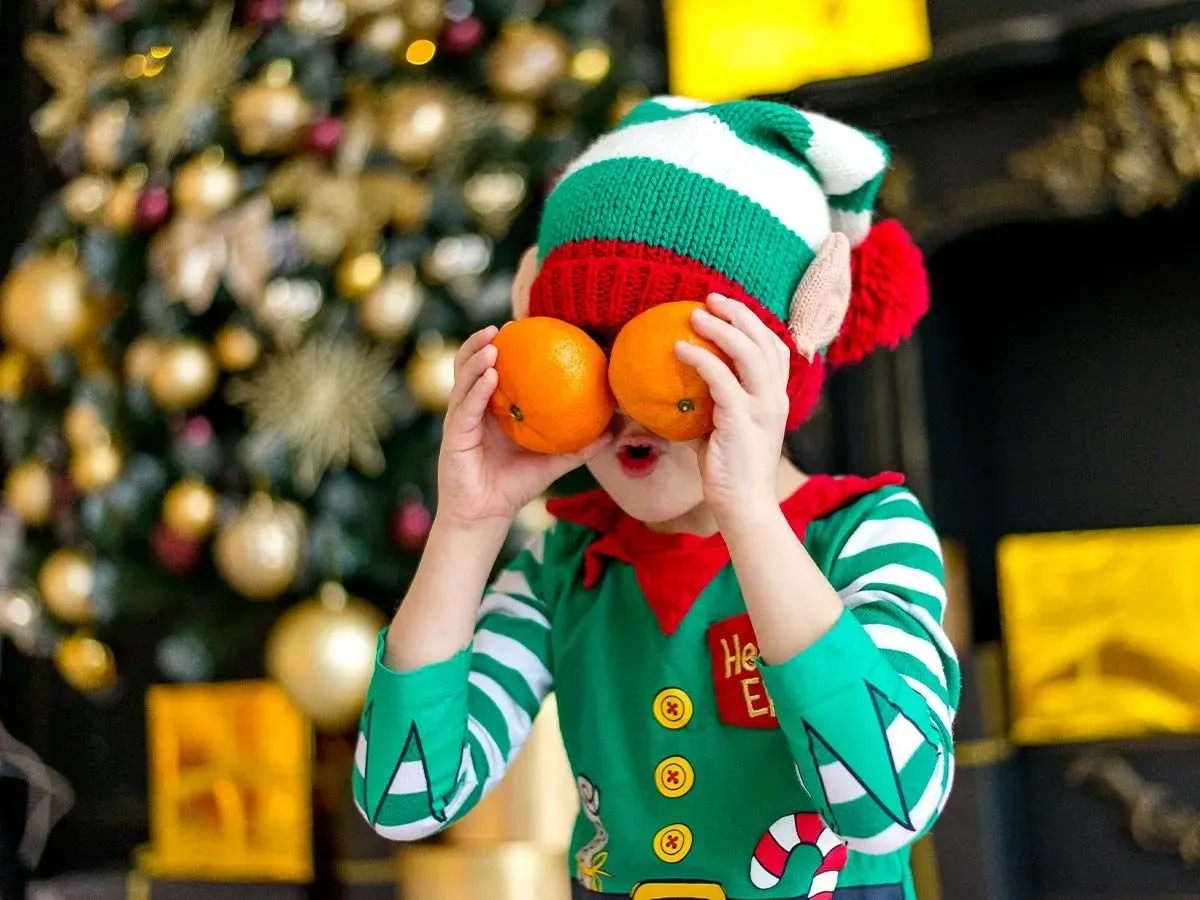 Child dressed as an elf making a silly face by holding up two oranges as googly eyes.