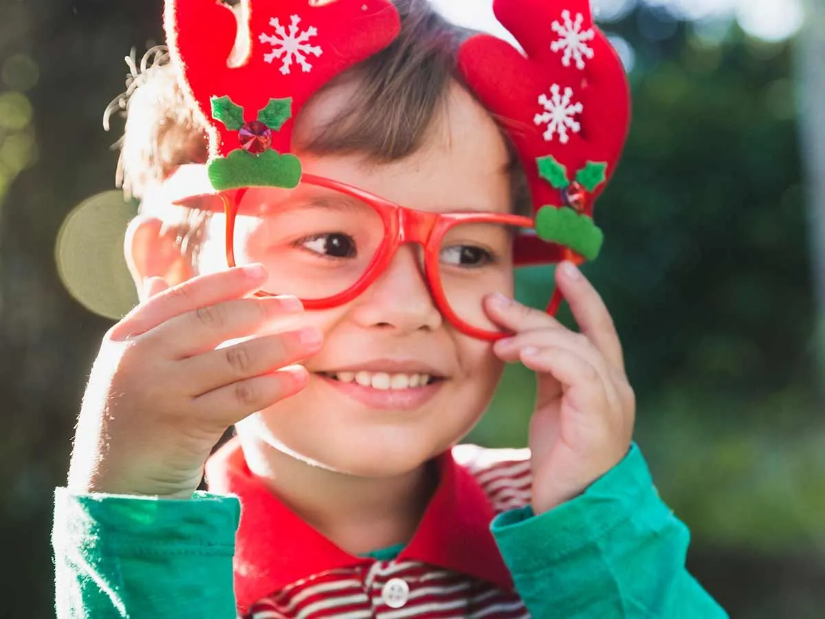 Young boy standing outside smiling, wearing red Christmas glasses with antlers ears on them.