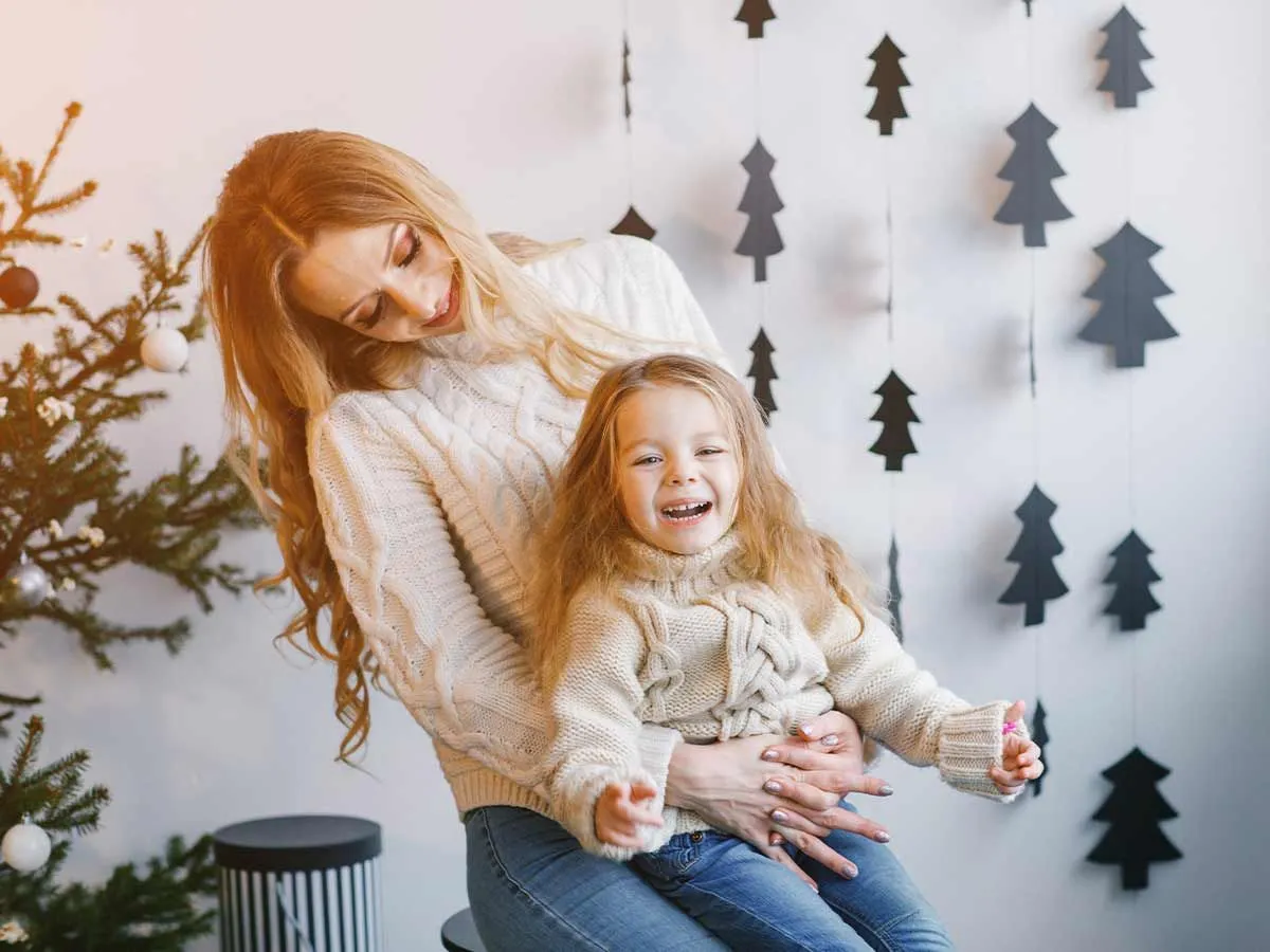 Mum and daughter laughing at reindeer jokes with Christmas tree decorations hanging behind.