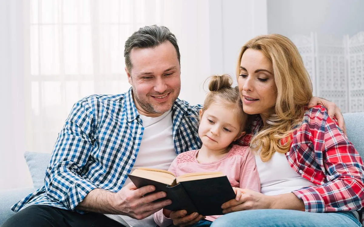 Parents sat on the sofa with their daughter reading a book to help her learn about the possessive apostrophe.