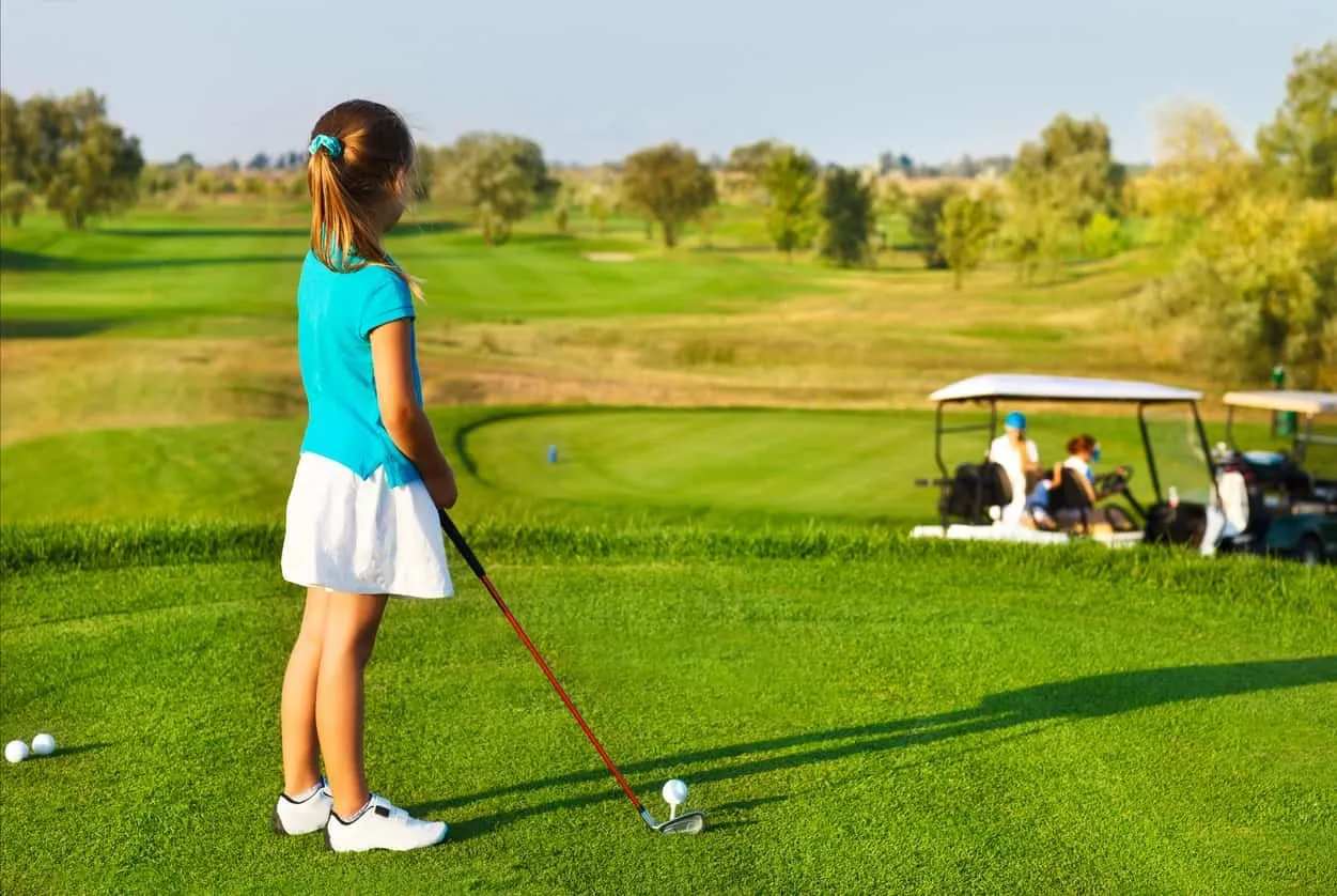 A young girl holding a kids' golf club stands with her back to the camera looking out over the golf course.