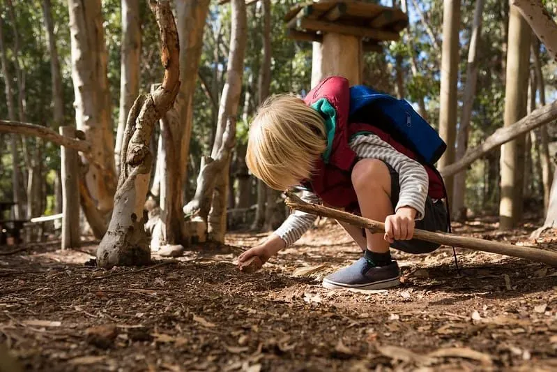 Little child with a backpack on their back crouching down inspecting a rock in the forest.