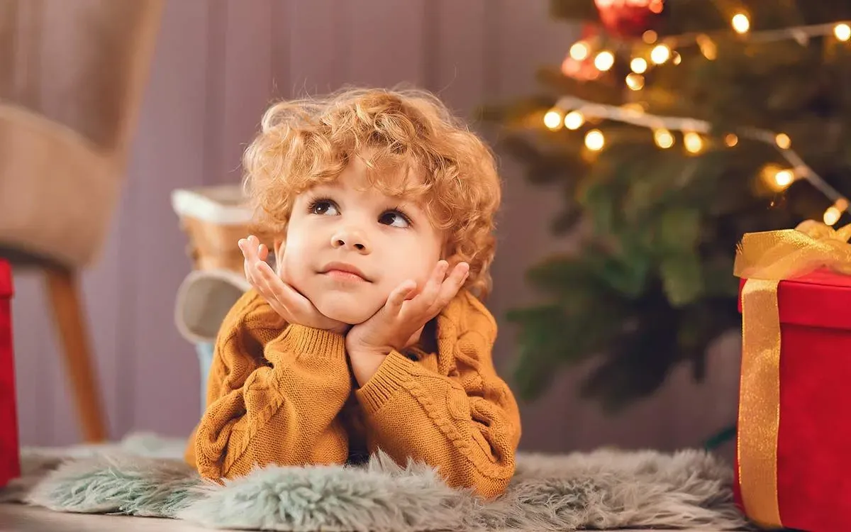 Little boy lying on his stomach by the base of the Christmas tree thinking about answers to Christmas riddles.