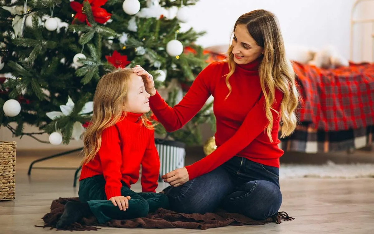 Mum and daughter sat on the floor by the Christmas tree smiling as they discuss Christmas riddles.
