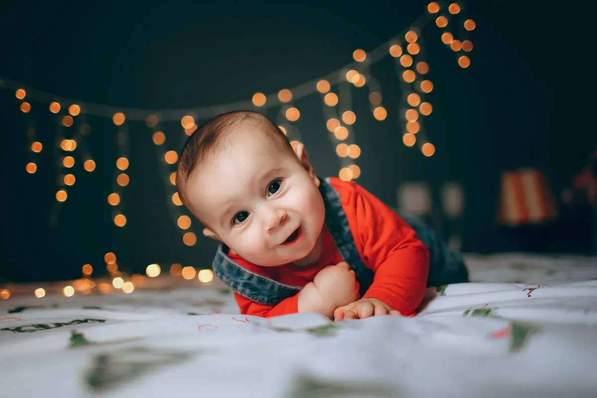 A baby boy is lying on his front on a bed looking at the camera, there are some magical fairy lights shining behind him.