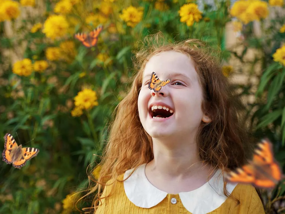 Girl standing by some plants in a garden with an orange butterfly on her nose, laughing at summer puns.