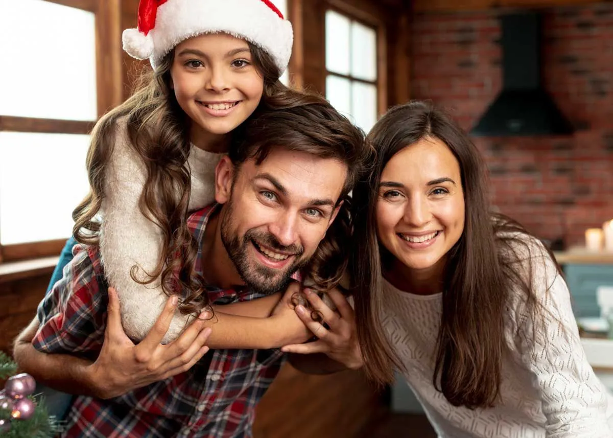 Young girl wearing a Santa hat hugging her dad while she smiles for a Christmas photo with her parents.