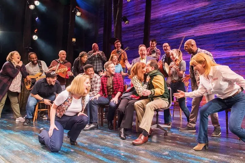 Two characters from Come From Away kissing on stage with the rest of the cast cheering around them.