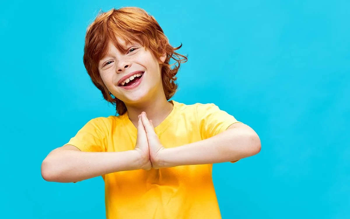 Young boy wearing a yellow t shirt smiling with his hands together standing in front of a blue background.