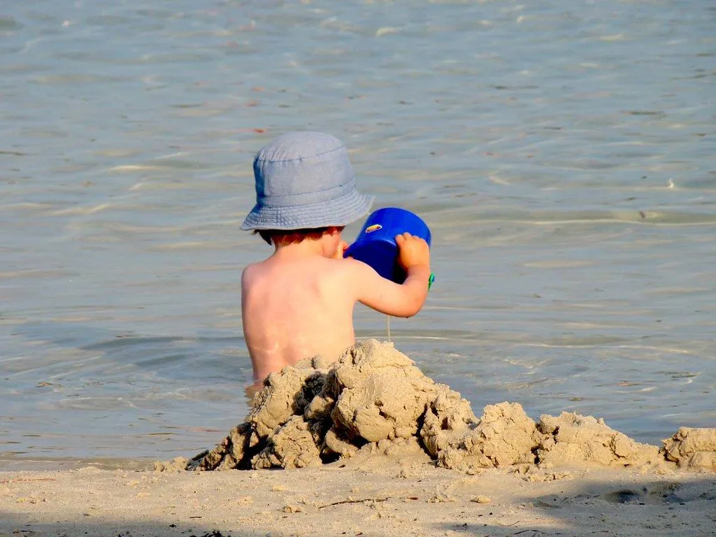 An image from behind of a young boy building a sandcastle and looking out to sea.a