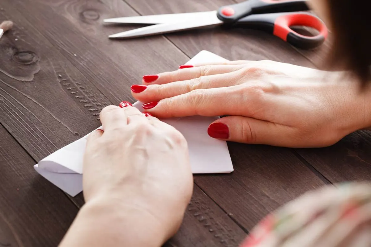 Close up of a woman with red nail polish folding a white piece of paper to make an origami sheep.