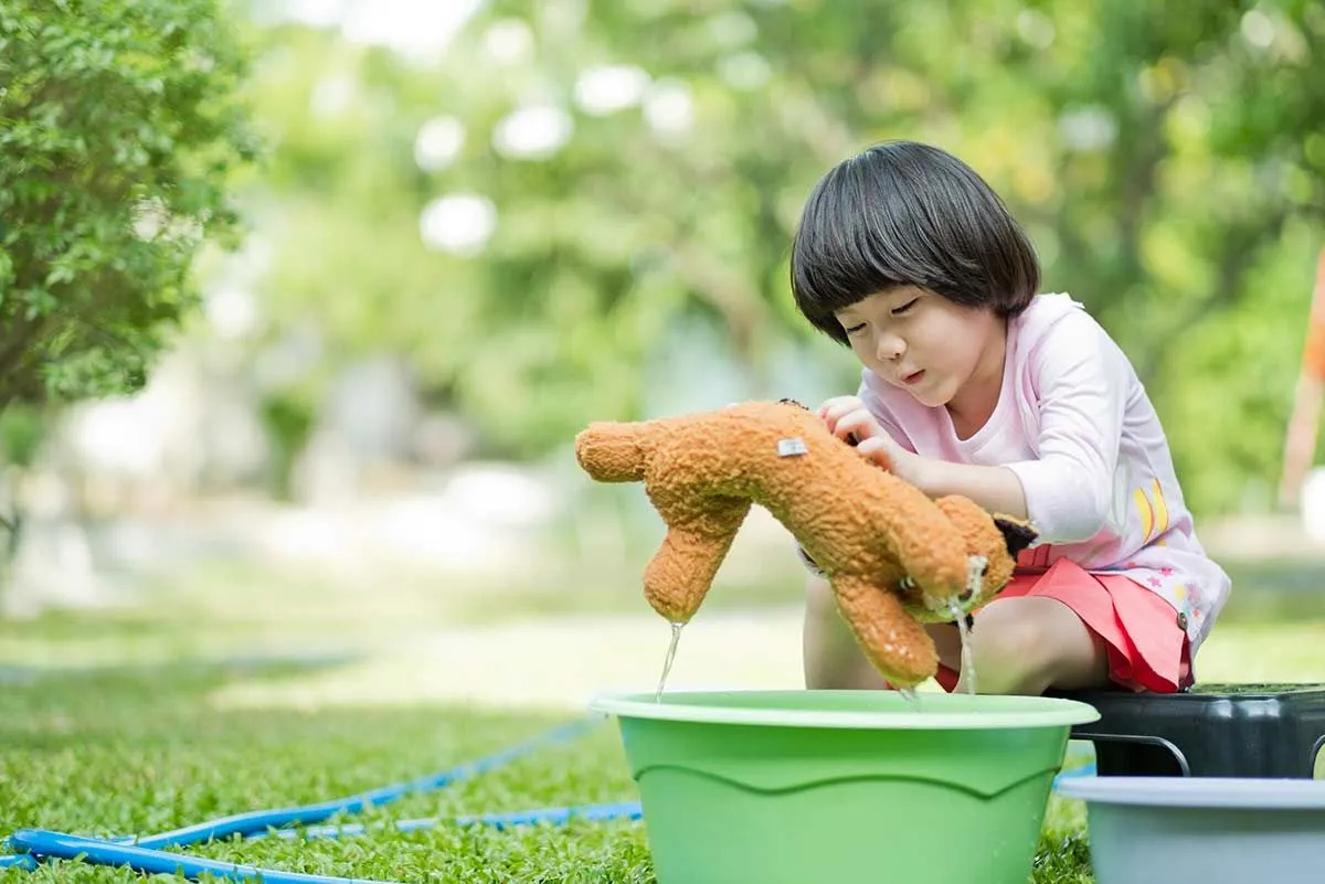 Little girl sat outside cleaning her teddy bear in a bucket of water.