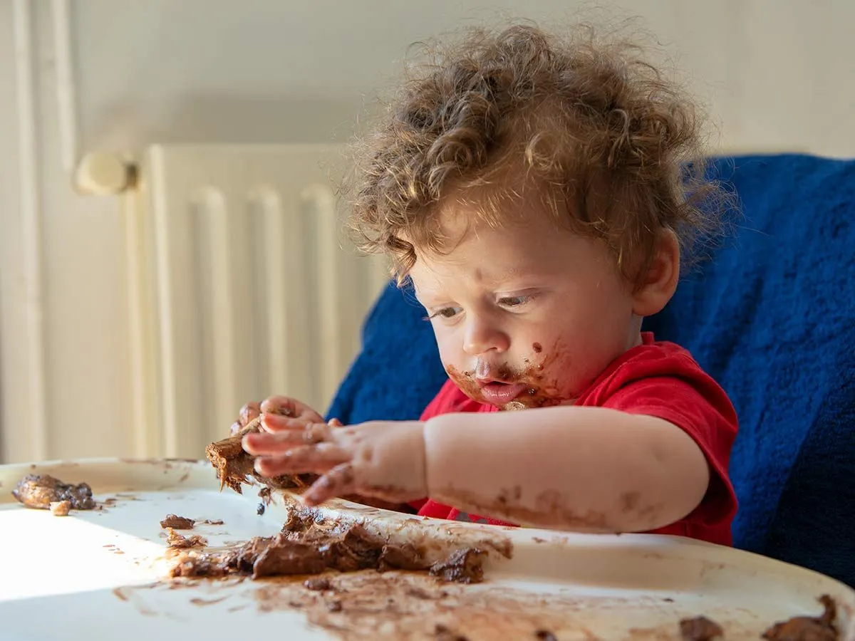 Young boy in a high chair eating a piece of chocolate monkey cake.