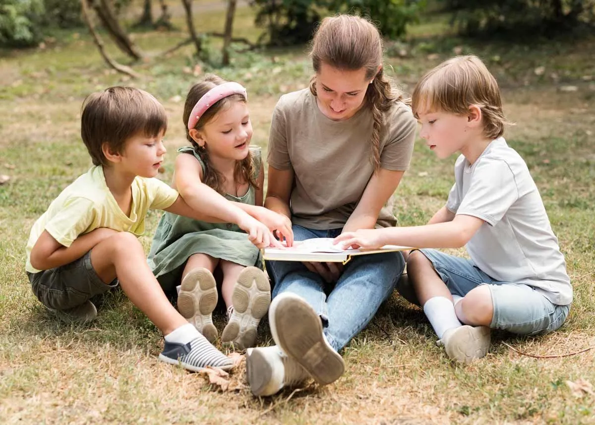 Mum and three kids sat on the grass outside reading a book together.