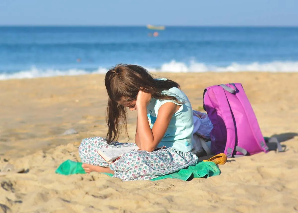 Little girl sat in the sand on the beach reading her book.