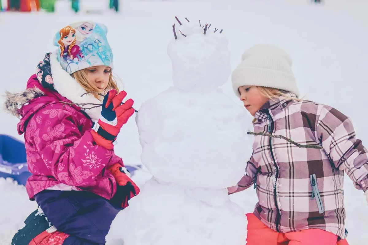 Two girls wrapped up in warm clothing playing outside in the snow building a snowman.