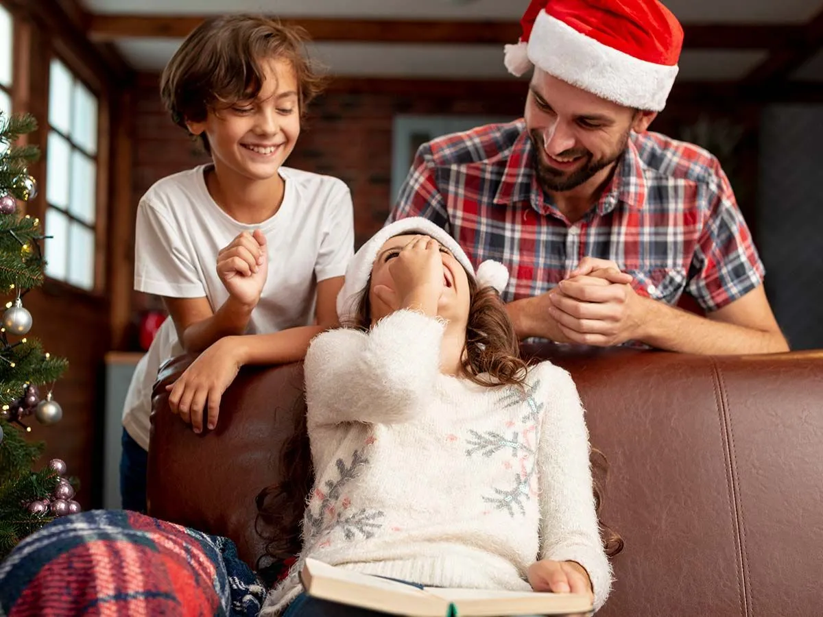 Dad and son standing behind the sofa, mum sat on the sofa laughing as they share Christmas jokes while wearing Santa hats.