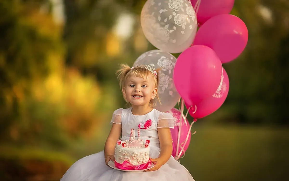 Little girl dressed in a pink princess outfit holding pink balloons in her garden at her party with family at home.
