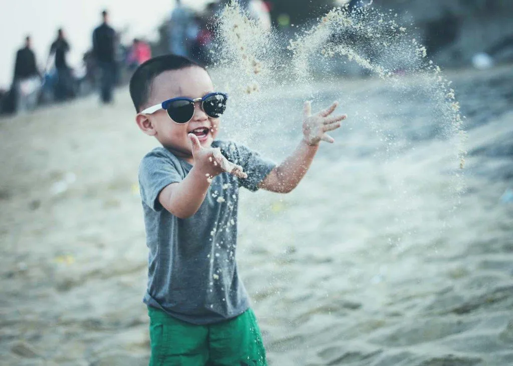 A little boy wearing sunglasses throws sand into the air at the beach.