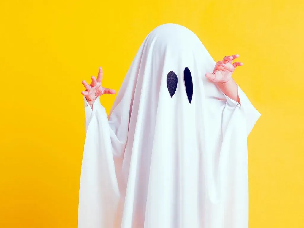 A child wearing a ghost costume poses in front of a yellow wall.