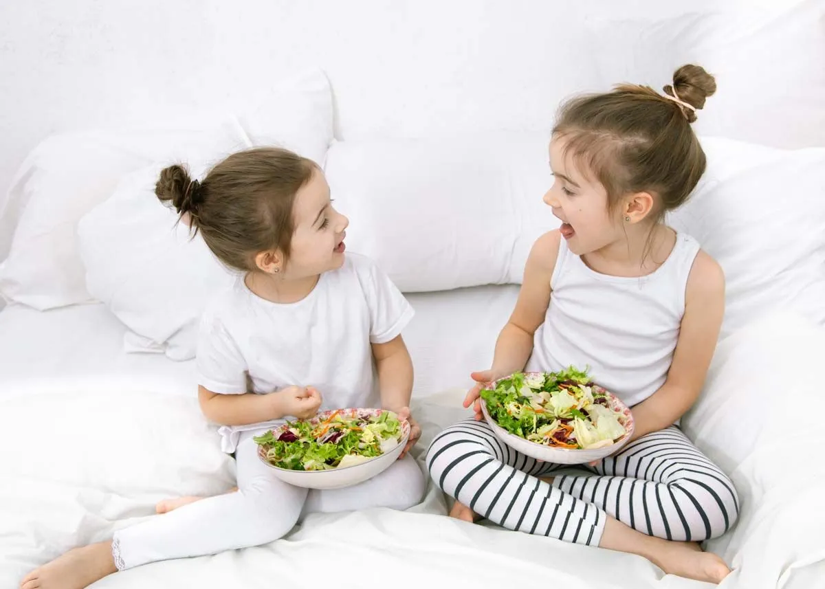 Two sisters sit on the bed smiling at each other whilst holding a bowl of salad in their hands.