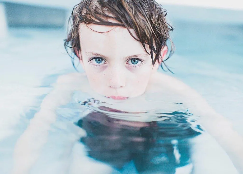 A little boy swimming in water looks directly at the camera.