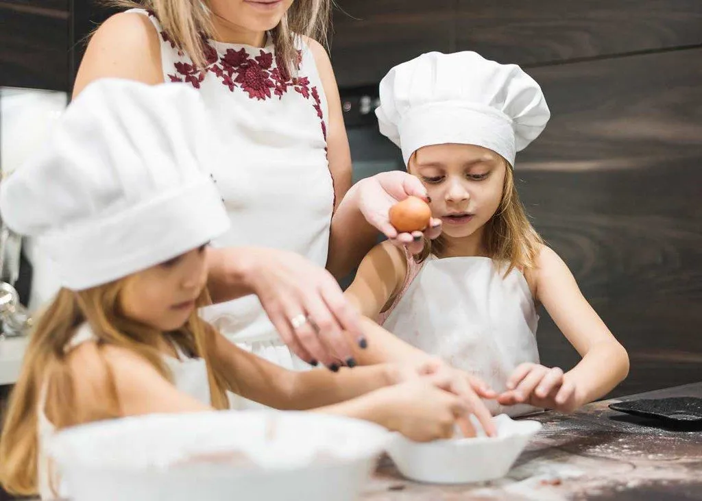 Two girls wearing chef hats are baking some food together with their mum.