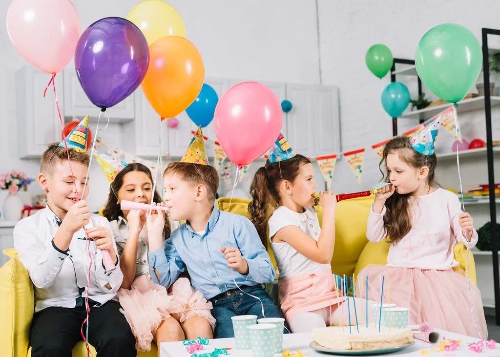 A group of children are sat amongst lots of balloons and other birthday party decorations.
