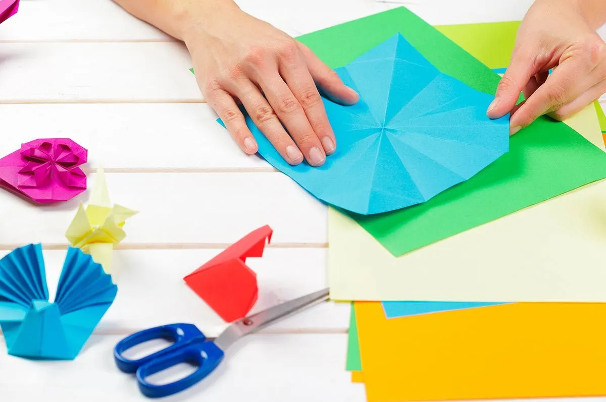 A close up image of a child's hands folding a blue sheet of origami paper to make an origami monkey.