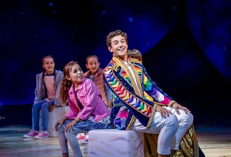 The main character Joseph in in the show Joseph and the Amazing Technicolor Dreamcoat wearing a colourful coat and sitting next to a group of child actors. 