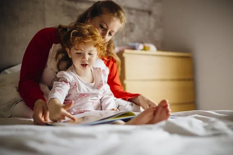 A mother reads a bedtime story to her little girl who is sat on her lap wearing a patterned onesie.