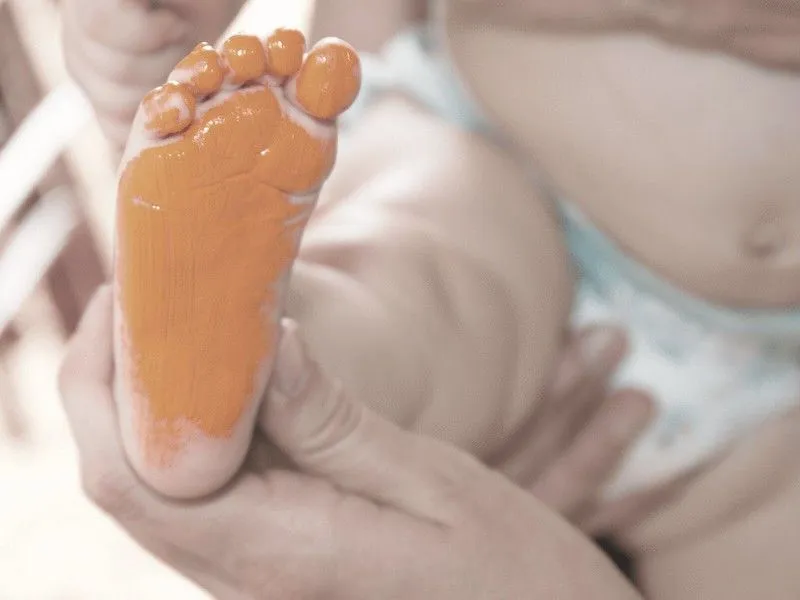 An adult's hands hold a tiny baby's foot which is covered in paint in order to make a baby footprint keepsake.