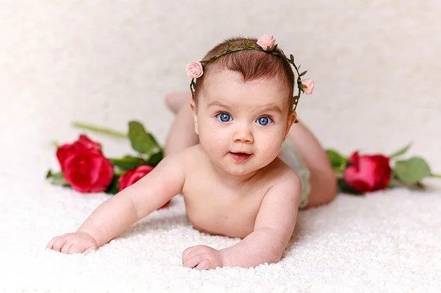 A newborn baby girl with blue eyes wearing pink roses headband is laying in the bed of red roses
