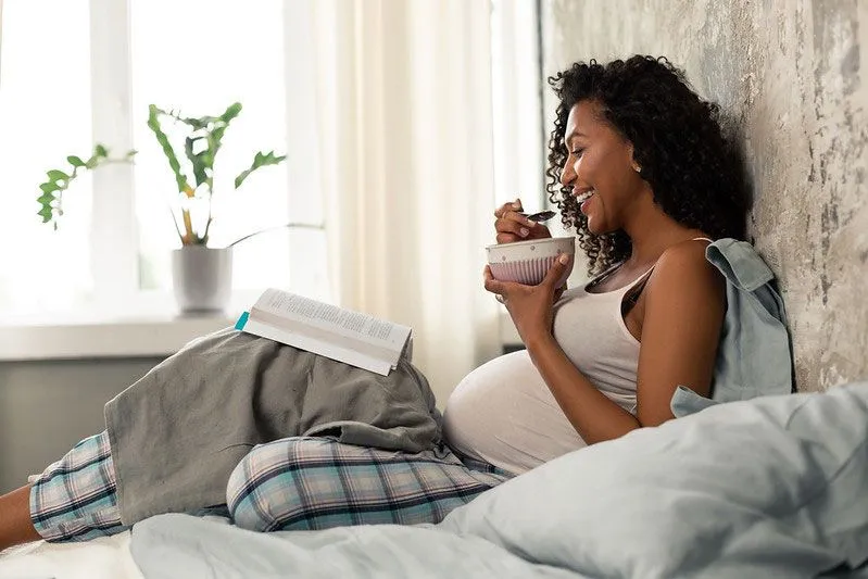 Pregnant woman laying in bed eating cereal and reading